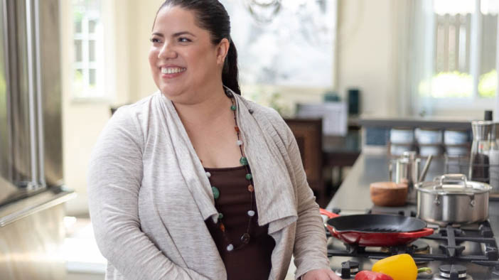 Smiling woman standing in a kitchen, joyfully preparing food with vegetables and pots on the countertop.