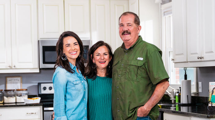 Man smiling alongside his wife and daughter in a kitchen, posing for a group photo.