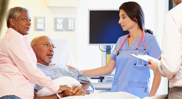 Patient in a hospital bed talking to a nurse, with a caregiver by his side