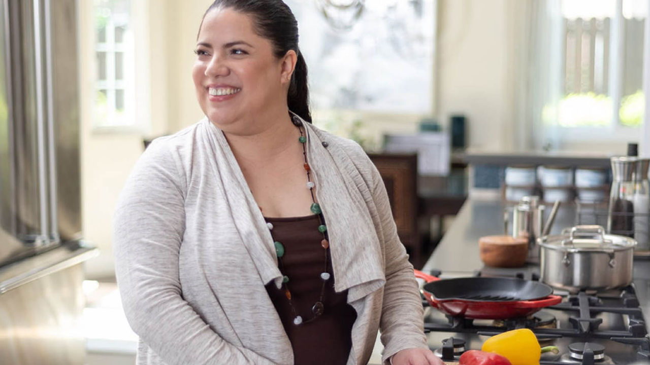 Smiling woman standing in a kitchen, joyfully preparing food with vegetables and pots on the countertop.