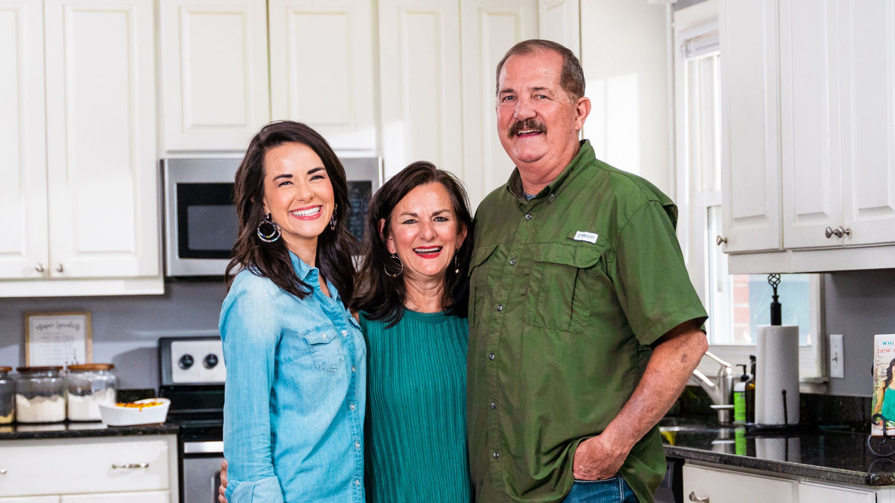 Man smiling alongside his wife and daughter in a kitchen, posing for a group photo.